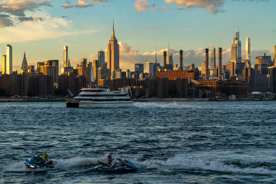 A ferry and two jet skis on the river below the Midtown skyline at golden hour