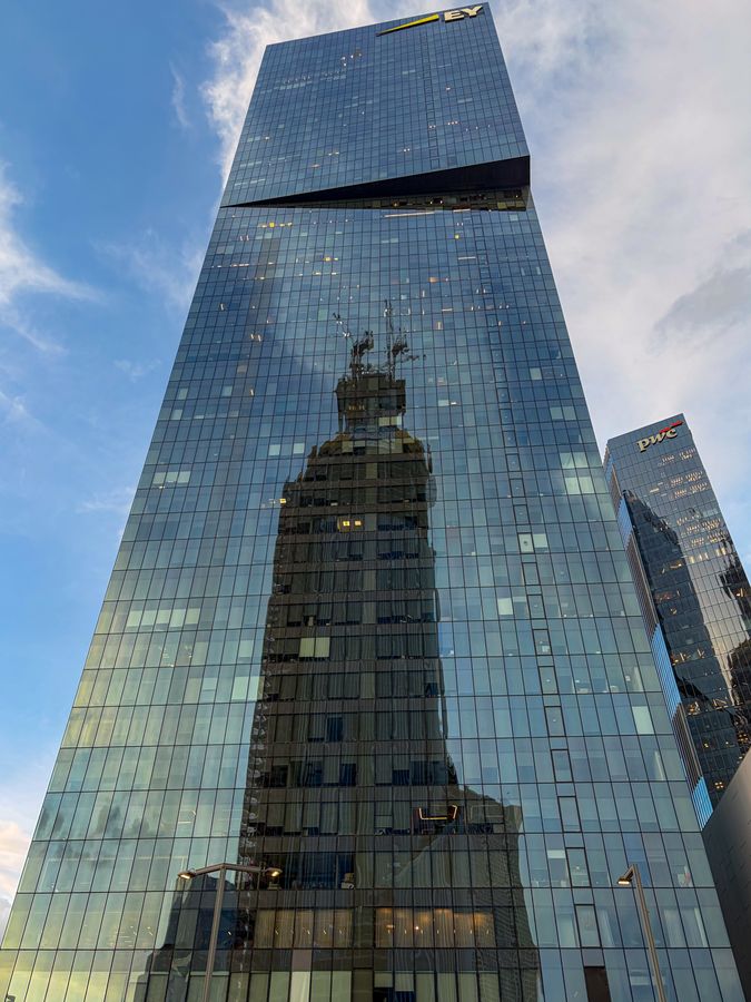 A glass tower reflecting another building against a blue sky